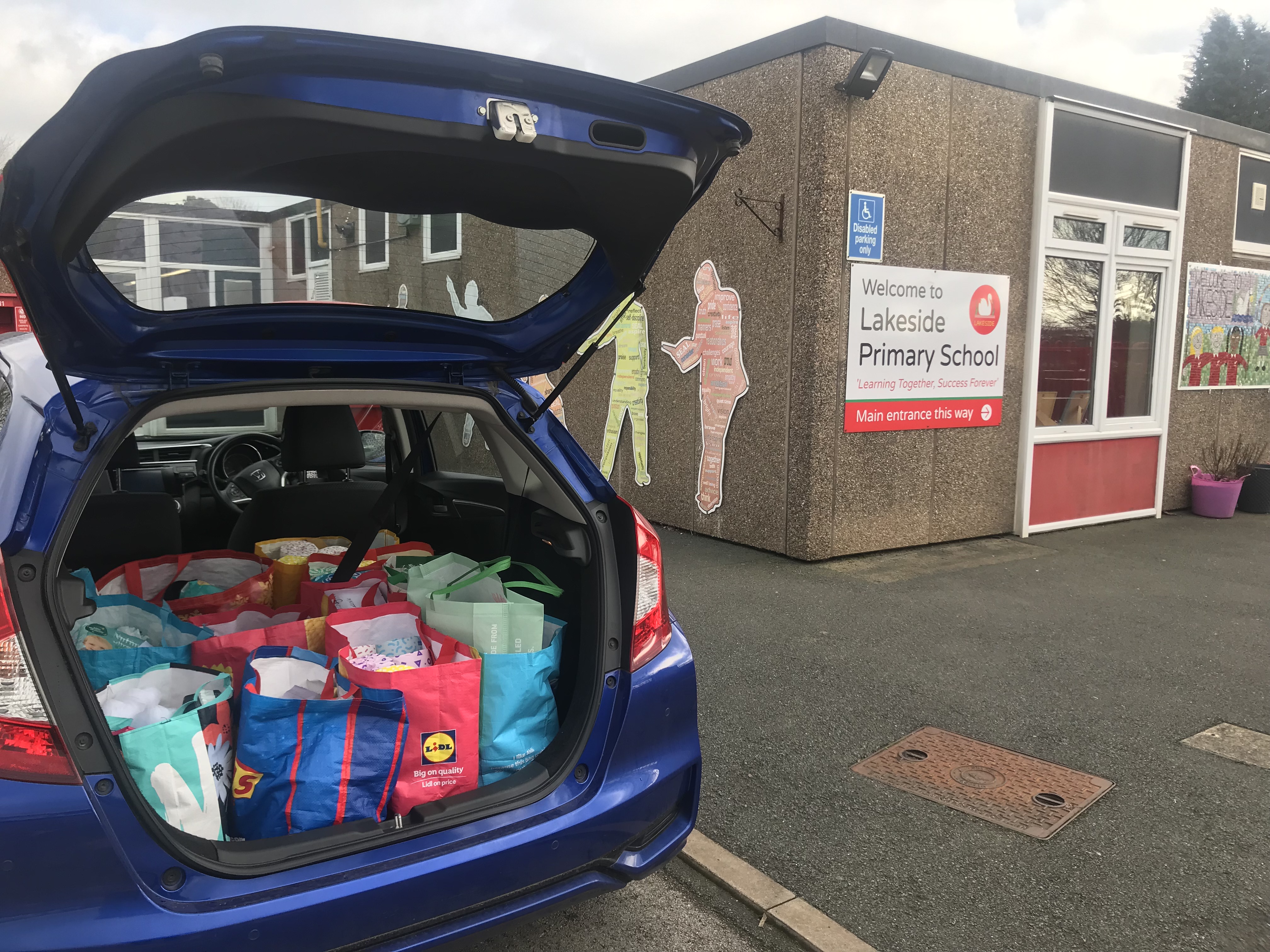 A car with its boot open with lots of bags full of hygiene products parked next to a building