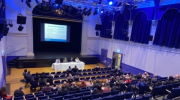 An auditorium with chairs and people sat listening to four people sat at a table at the front
