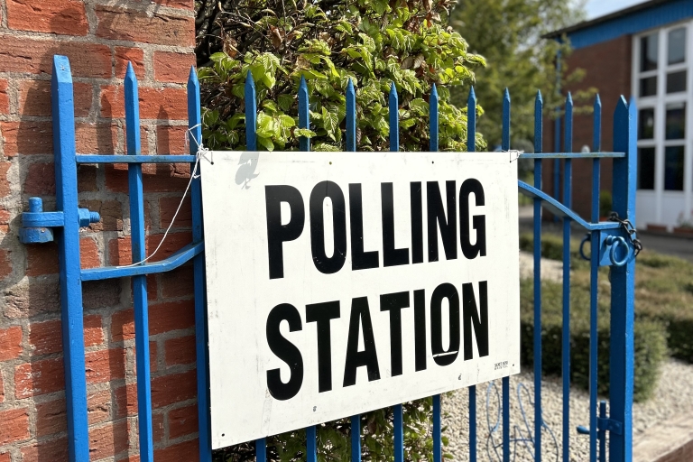 'polling station' sign on a blue fence next to a brick wall and a tree