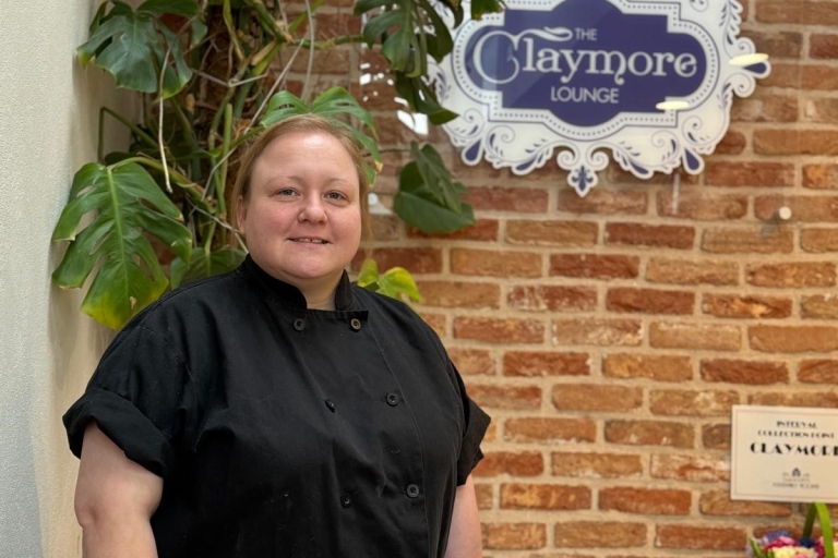 photo of a chef in black jacket standing by a table and brick wall in the claymore lounge