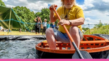 A man with a white beard in a yellow top and hat sat on a coracle boat with an oar in his hand on some water. There are people in the background watching on a blue sky, cloudy day.