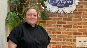 photo of a chef in black jacket standing by a table and brick wall in the claymore lounge