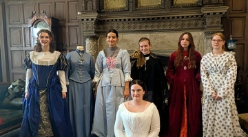 people sitting and standing in Victorian clothes by a fireplace and wooden panel walls