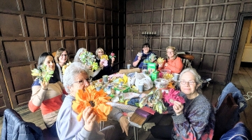 photo of a group of people in a room with dark wooden panelling, holding handmade material and wool flowers